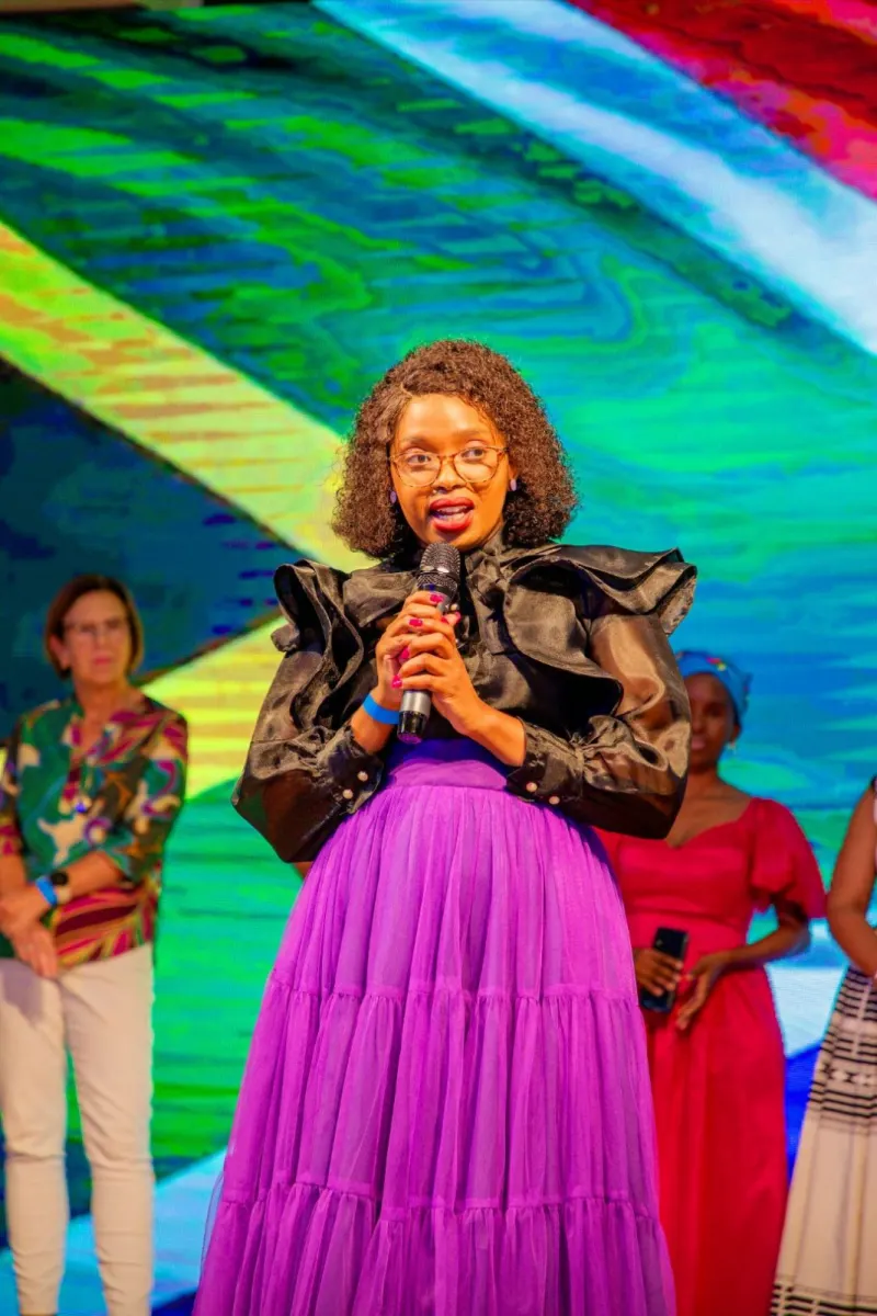 Woman in black ruffled top and purple skirt speaks into microphone on colorful stage