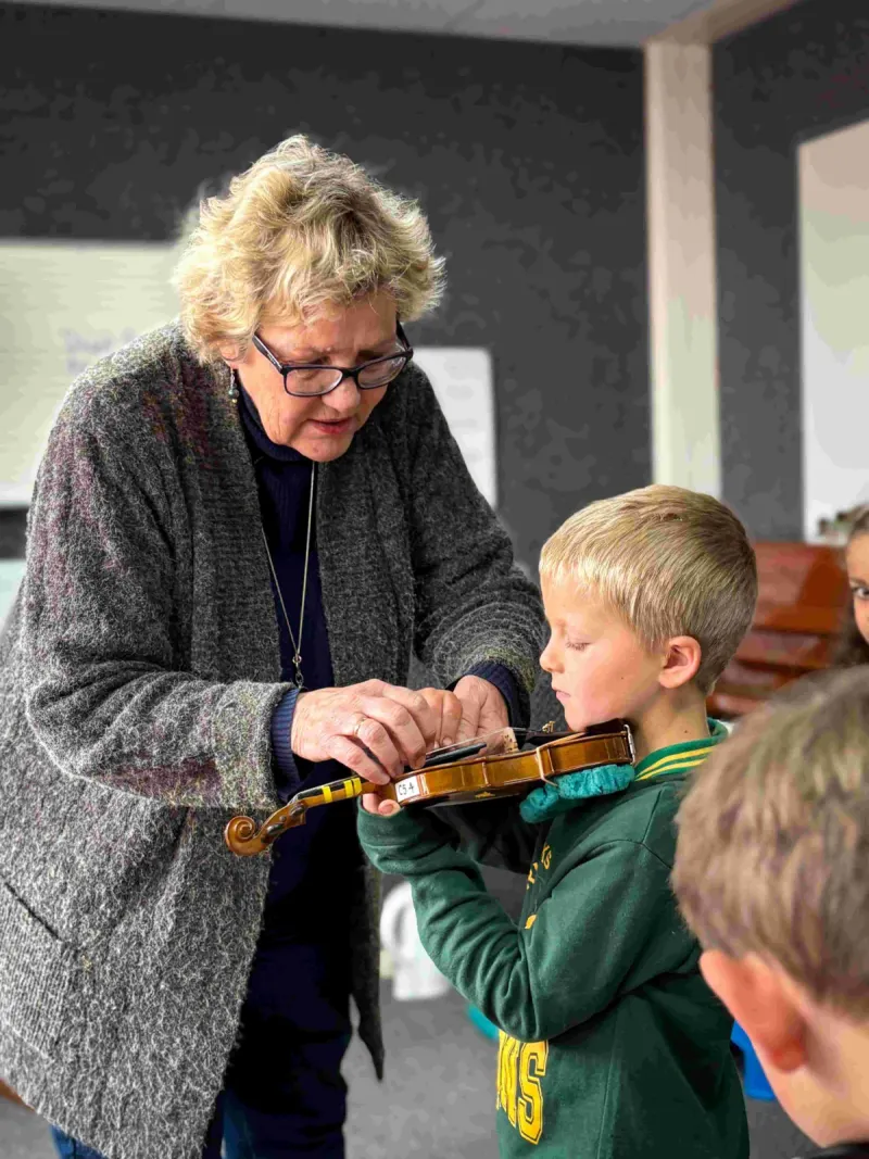 Older woman with glasses helping young boy hold a violin properly